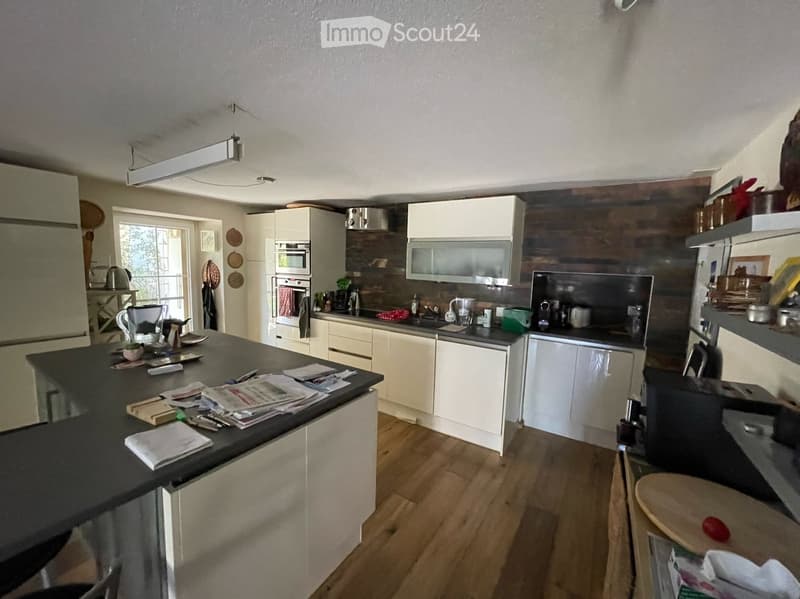 Modern kitchen with white cabinets, wooden accents, and a black countertop. Appliances include a refrigerator, oven, microwave, and dishwasher. The space features a central island with a stool and various kitchen items on the counter.
