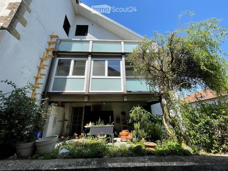 A modern house with a raised ground floor, large windows, and a well-maintained garden area with potted plants and greenery.