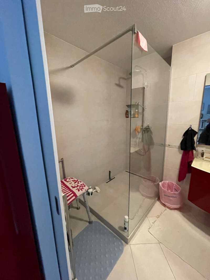Modern bathroom with a glass-enclosed shower stall, a towel hanging on the door, a mirror, a sink, and a cabinet. The floor is tiled, and there is a blue bath mat inside the shower area.