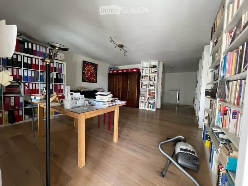 A well-organized study or office space with wooden flooring, a large wooden table with a laptop, books, and papers, multiple bookshelves filled with books and binders, a tall floor lamp, a wooden cabinet, and a vacuum cleaner on the floor.