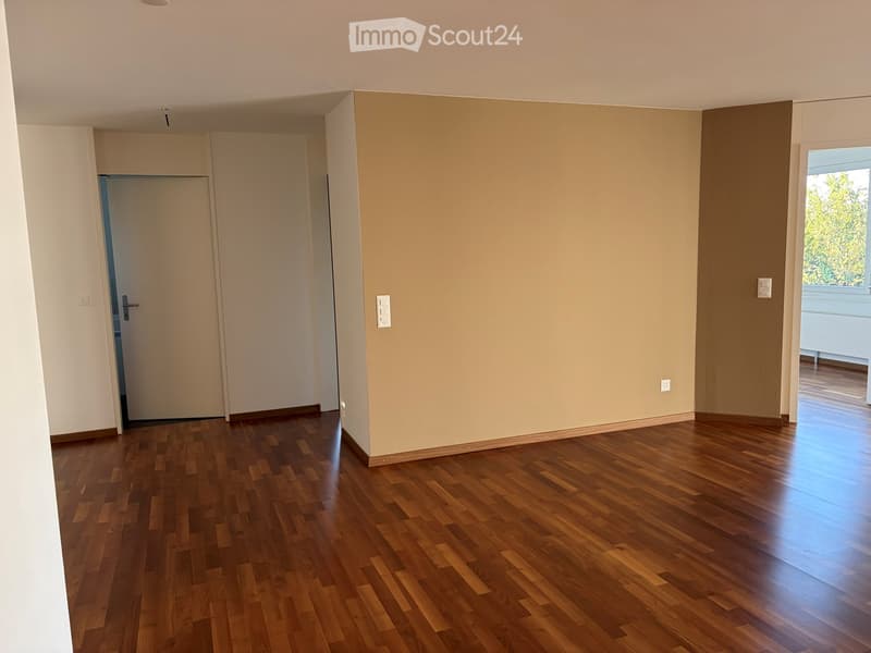 Empty room with wooden flooring, white walls, and a door leading to another room. Natural light comes through a window on the right side.