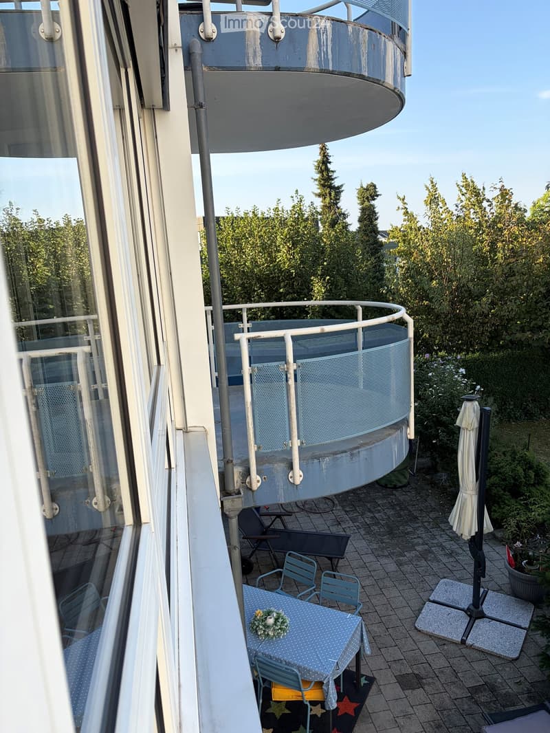 Balcony with a round seating area, a small table with a blue polka dot tablecloth, and several chairs. There is an umbrella and a view of greenery.