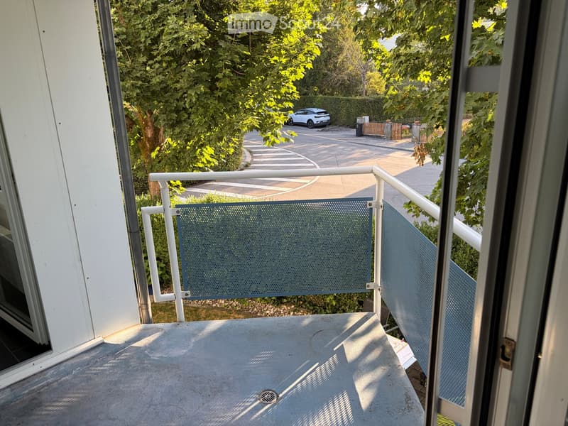 Balcony with white railings, metal mesh panels, and a view of a parking area with a car and greenery.
