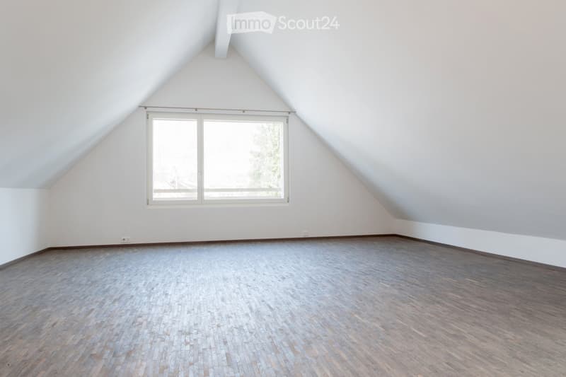 Empty attic room with wooden flooring, white walls, and a large window with a view of trees.