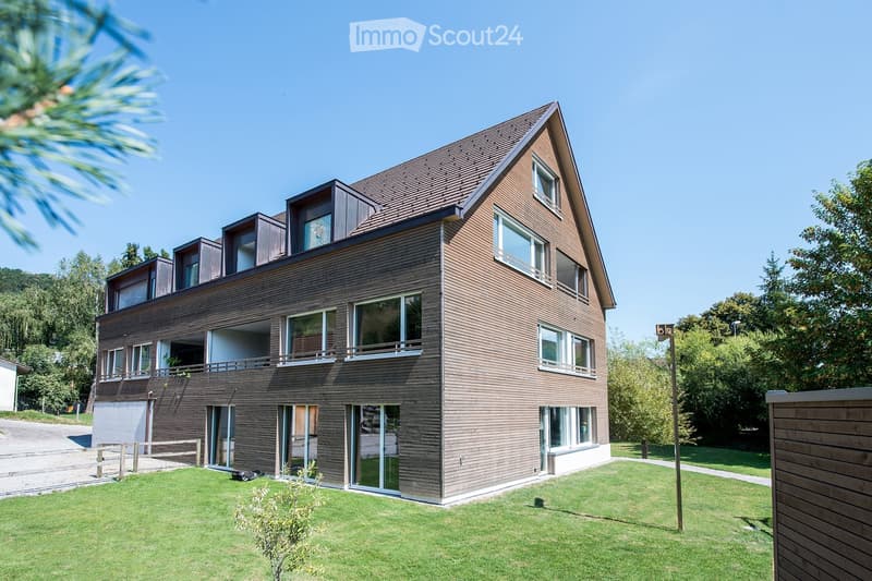 Modern two-story house with wooden siding, large windows, and a sloped roof. Features include a raised ground floor, multiple balconies, and a well-maintained lawn.