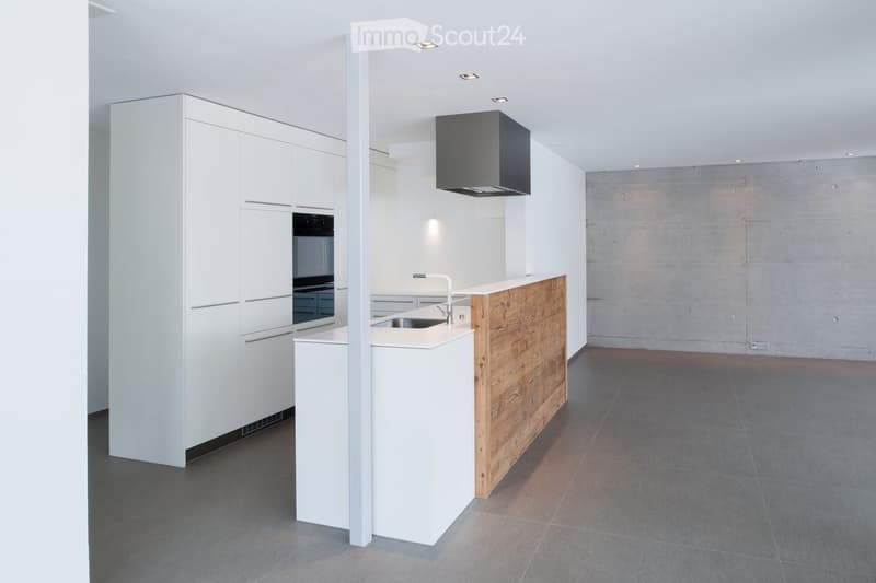 Modern kitchen with white cabinets, a central island with a wooden accent, a sink, and an oven. The space features a sleek, minimalist design with recessed lighting and a concrete accent wall.