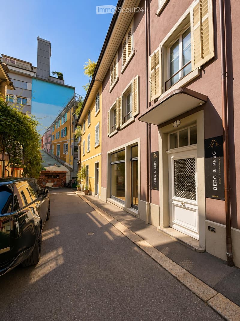 A narrow street lined with multi-story buildings featuring classic European architecture, including shutters and decorative facades. A parked car is visible on the left side.