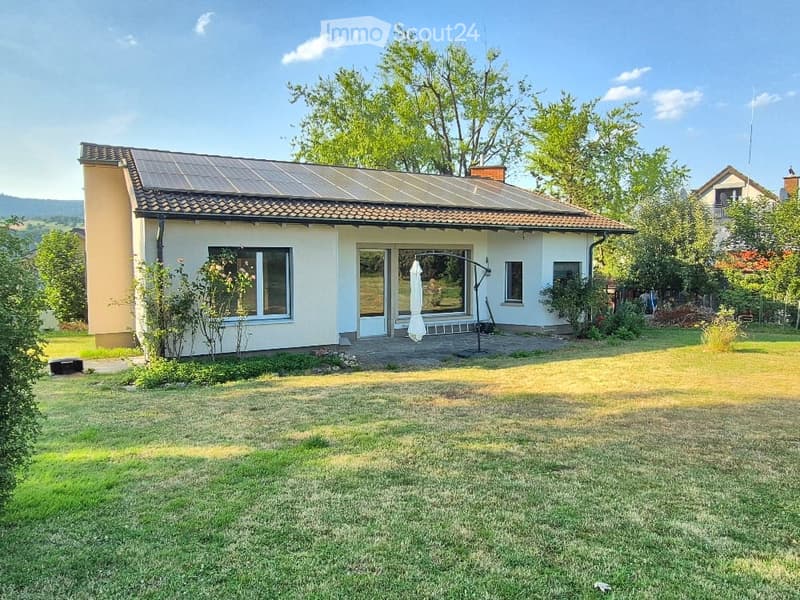 Single-story house with a tiled roof, large windows, and a covered patio area. The house is surrounded by a well-maintained lawn and trees.