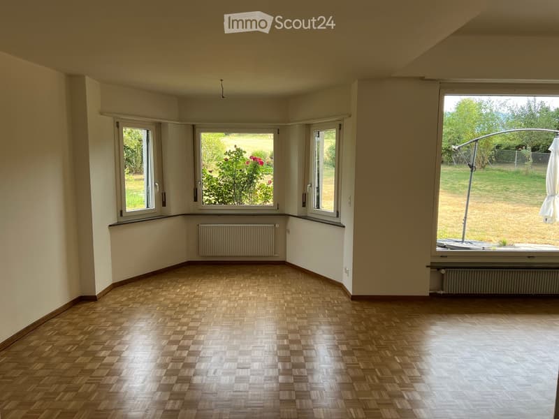 Empty room with wooden parquet flooring, large windows, radiators, and a view of a garden.