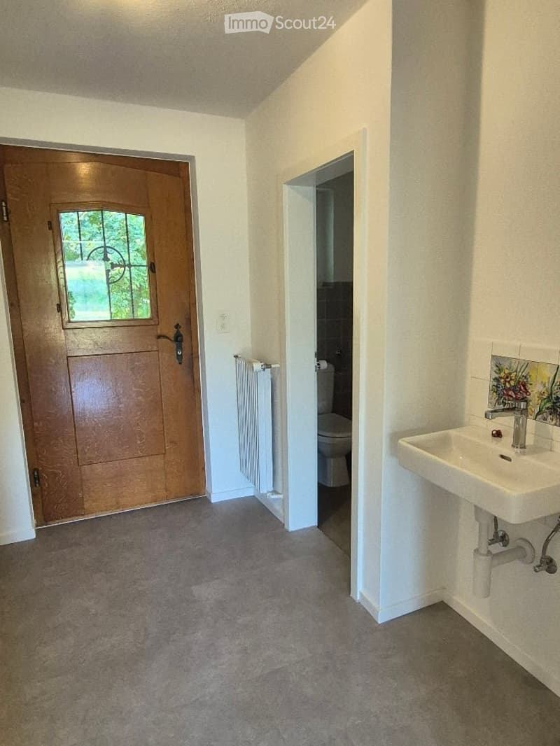 Empty hallway with a wooden front door, a radiator, and an open doorway leading to a bathroom with a sink and toilet.