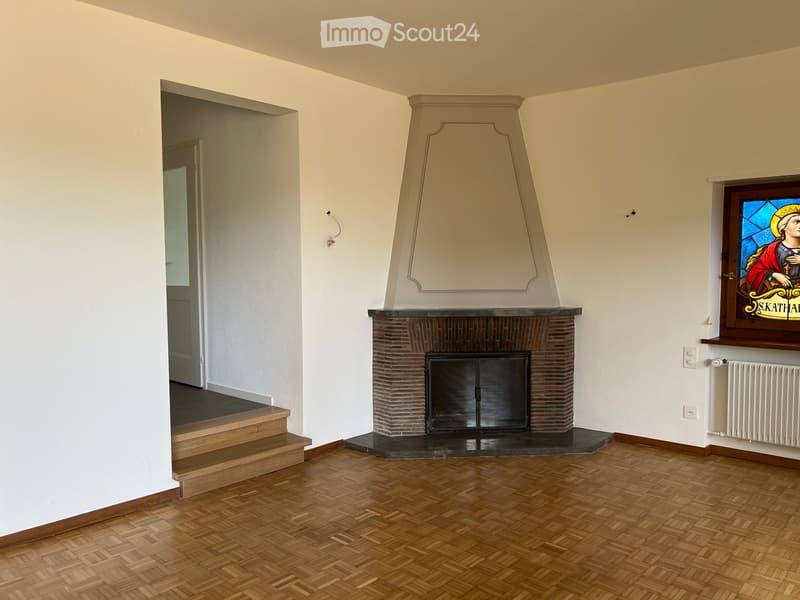 Empty room with wooden herringbone flooring, white walls, a brick fireplace with a decorative mantel, and a stained-glass window above it. A staircase leads to another area.