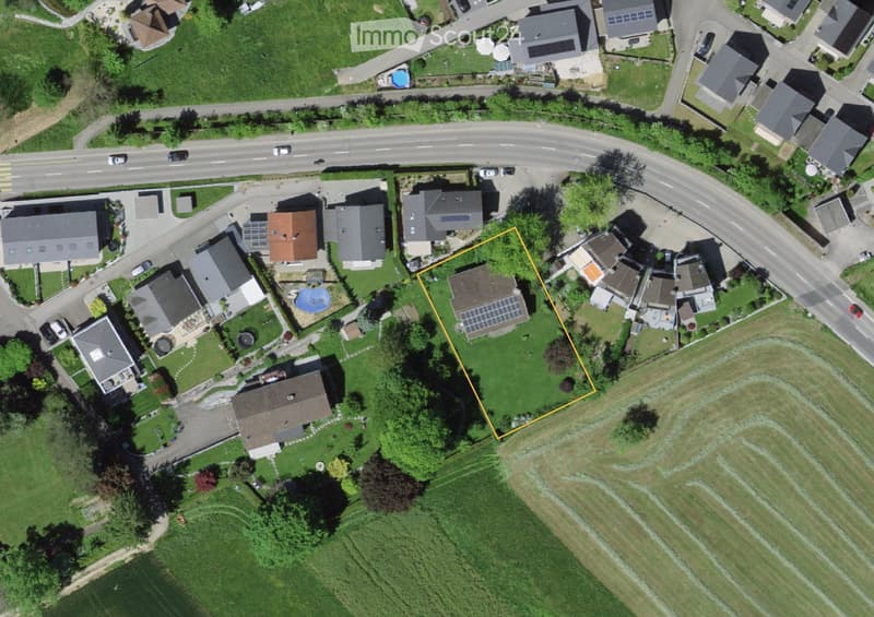 Aerial view of a residential area with multiple houses, gardens, and a road. The highlighted area shows a green lawn with trees and a small structure, likely a shed or garage.