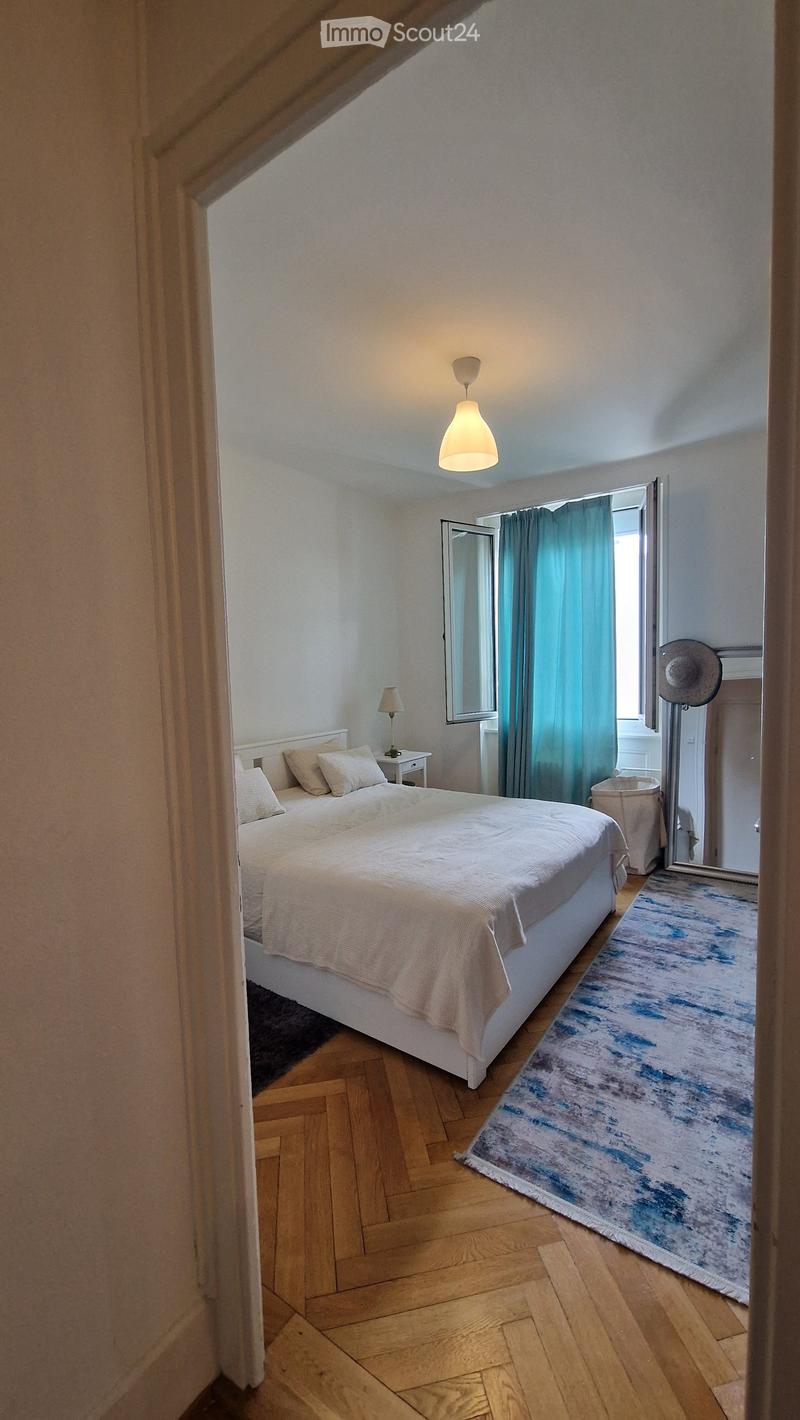 Cozy bedroom with a white bed, nightstand, lamp, mirror, and a blue and white rug on a wooden floor.