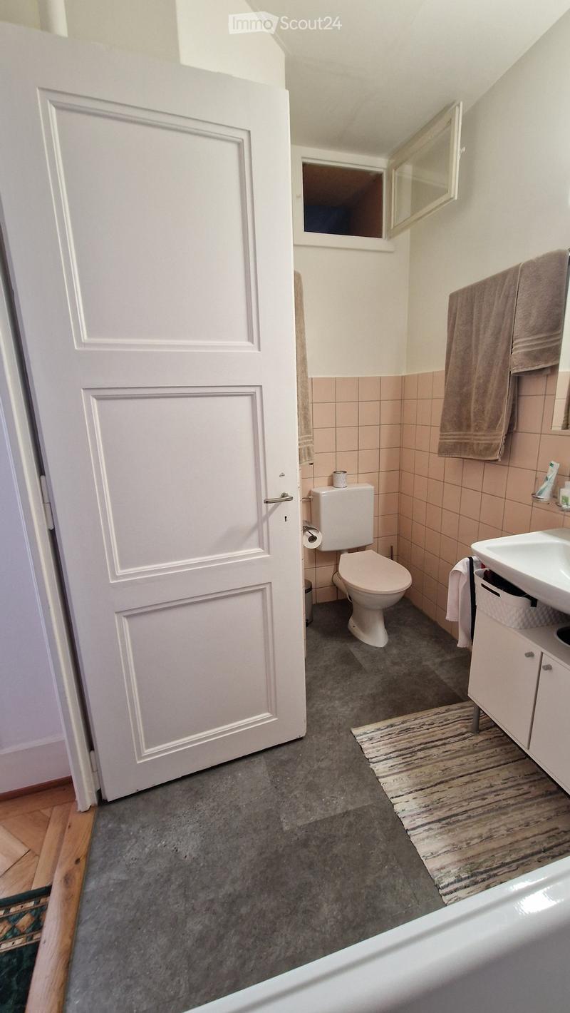 Modern bathroom with a toilet, sink, mirror, and towel rack. The room features light pink tiles, a grey floor, and a small rug near the sink.