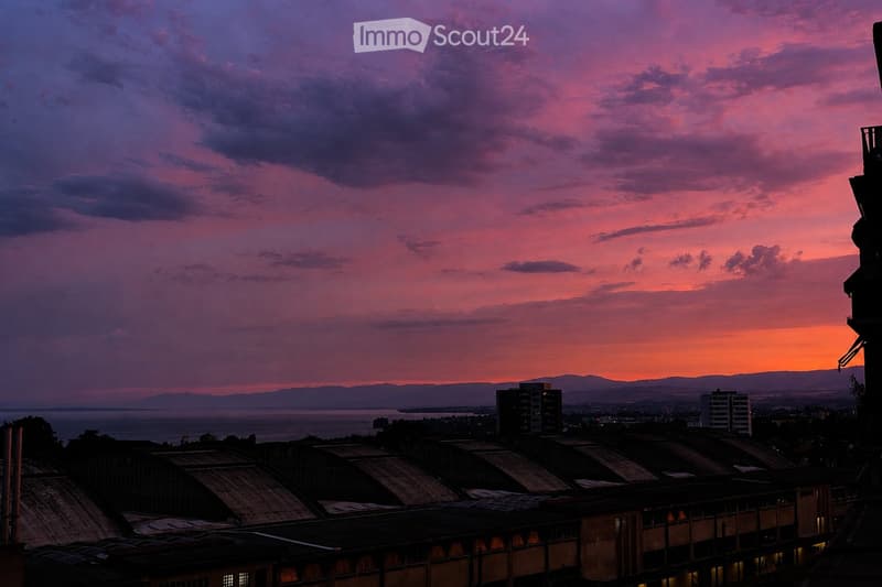 A panoramic view from a rooftop or balcony showcasing a vibrant sunset over a cityscape with distant mountains and a body of water.