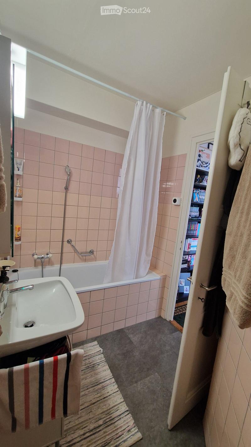 Bathroom with pink tiled walls, a bathtub with a shower curtain, a sink, a towel on a rack, and an open door leading to a bookshelf.