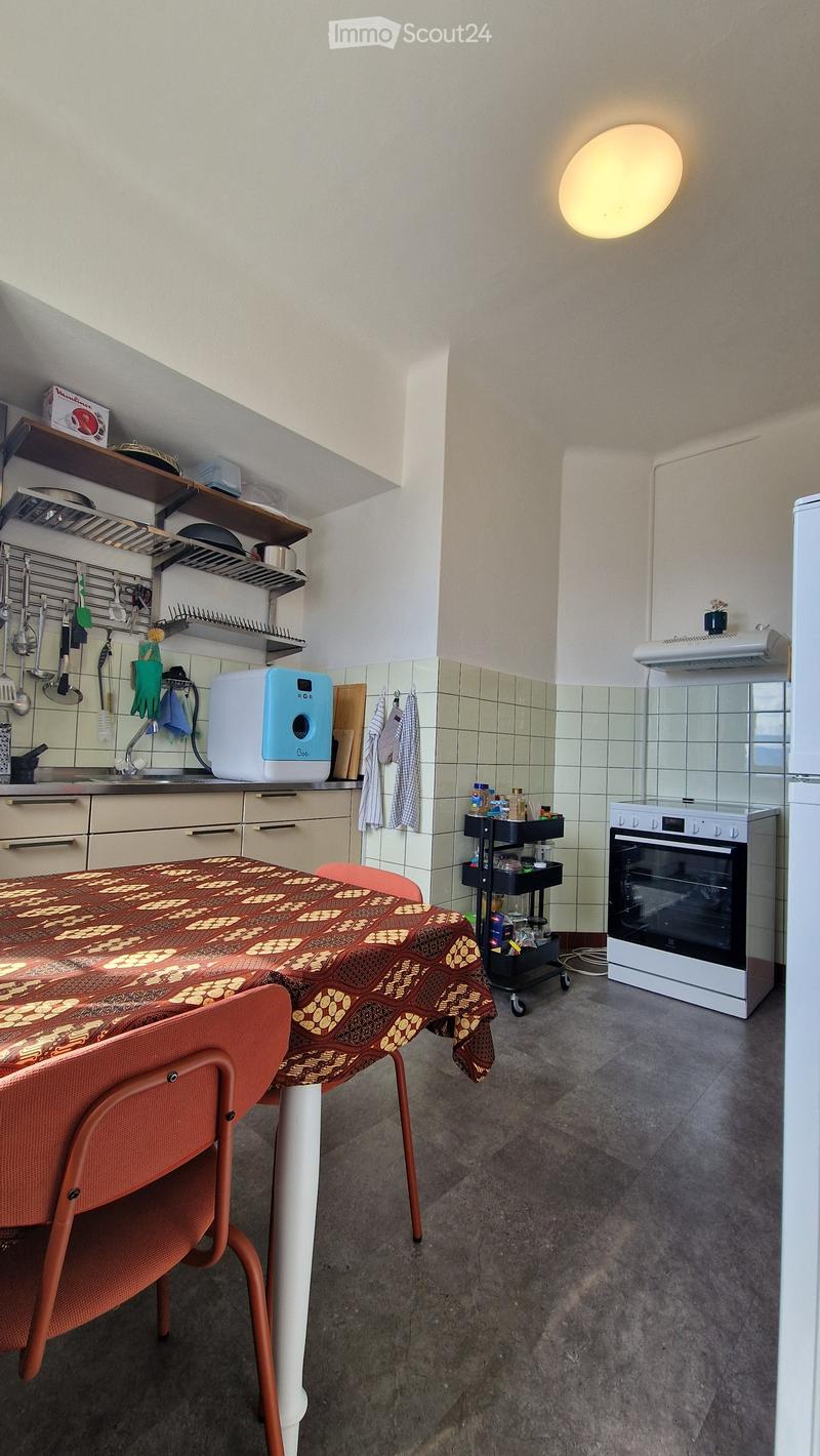 A modern kitchen with a dining area, featuring a table with a patterned tablecloth, red chairs, a white oven, a refrigerator, a dishwasher, and various kitchen utensils and appliances.