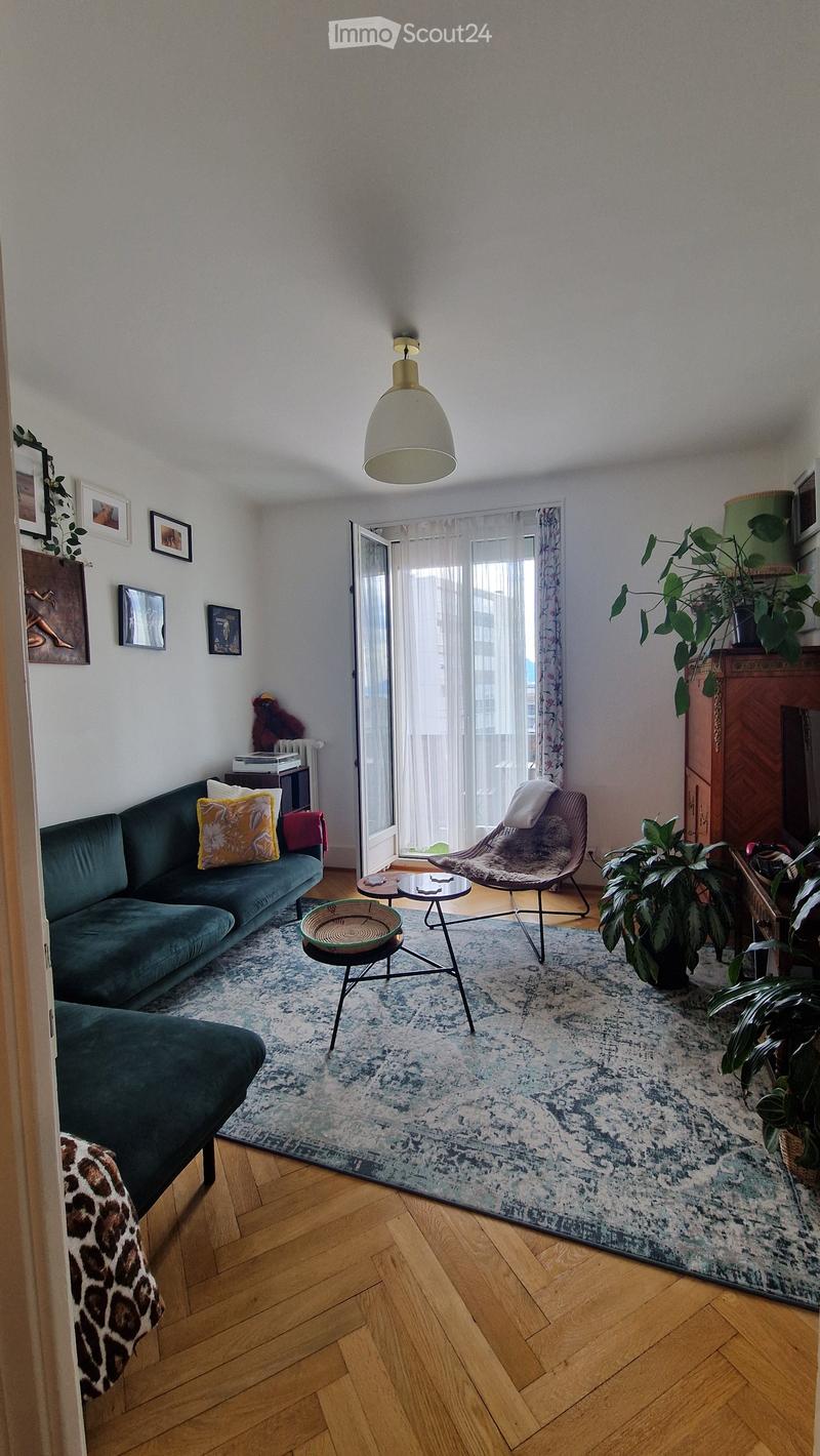 Living room with a green sofa, a wicker chair, a coffee table, a large rug, and several plants. The room features wooden flooring and large windows with sheer curtains.