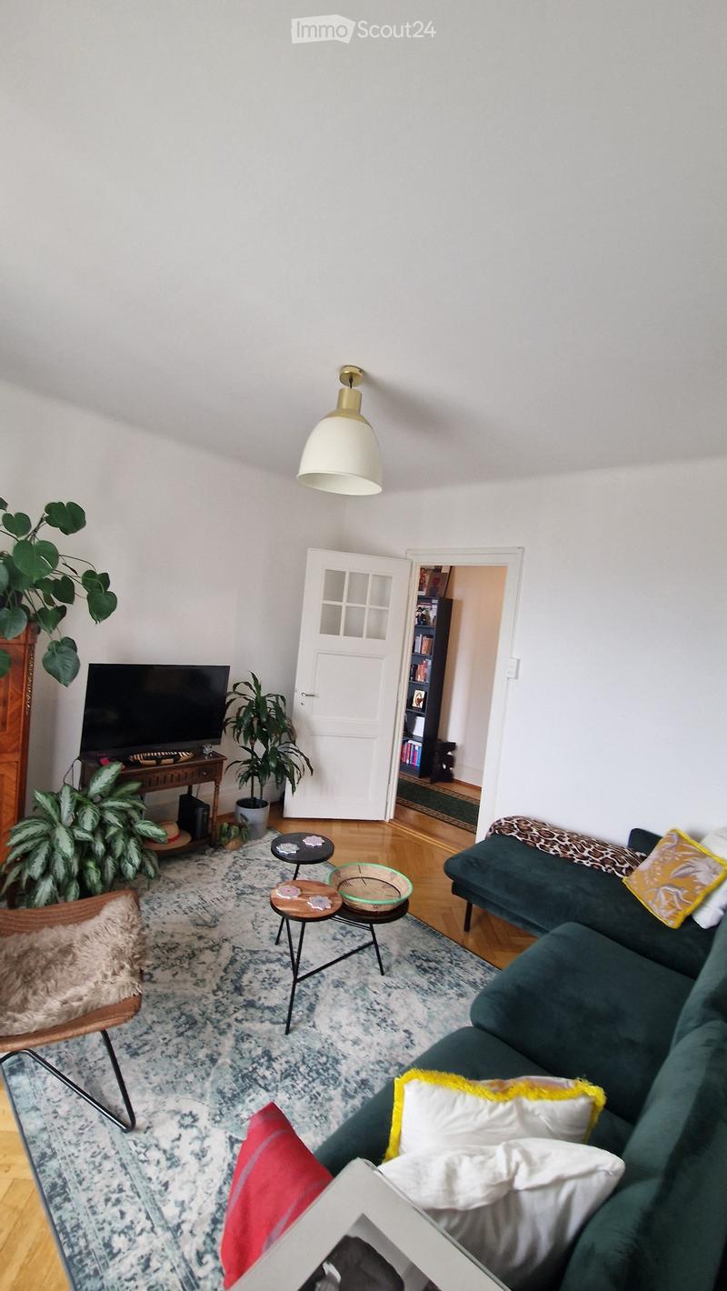 A modern living room with a green sofa, coffee table, small round tables, a hanging lamp, and a television on a wooden stand. The room features a patterned rug, wooden flooring, and several potted plants.