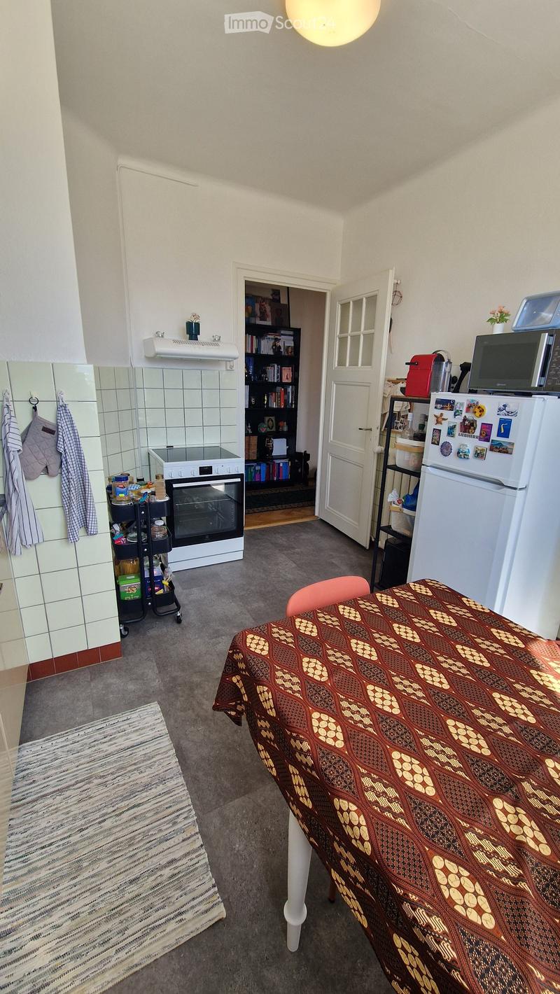 A small, furnished room with a dining table, chair, refrigerator, microwave, oven, bookshelf, cart, and a towel hanging on the wall.