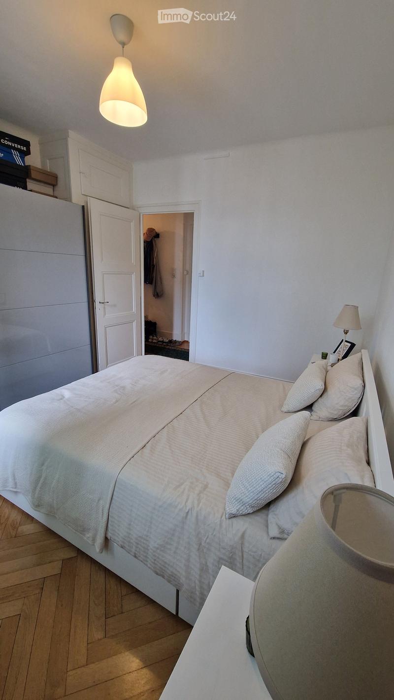 A modern bedroom with a neatly made bed, pillows, a nightstand with a lamp, and a wardrobe in the background.