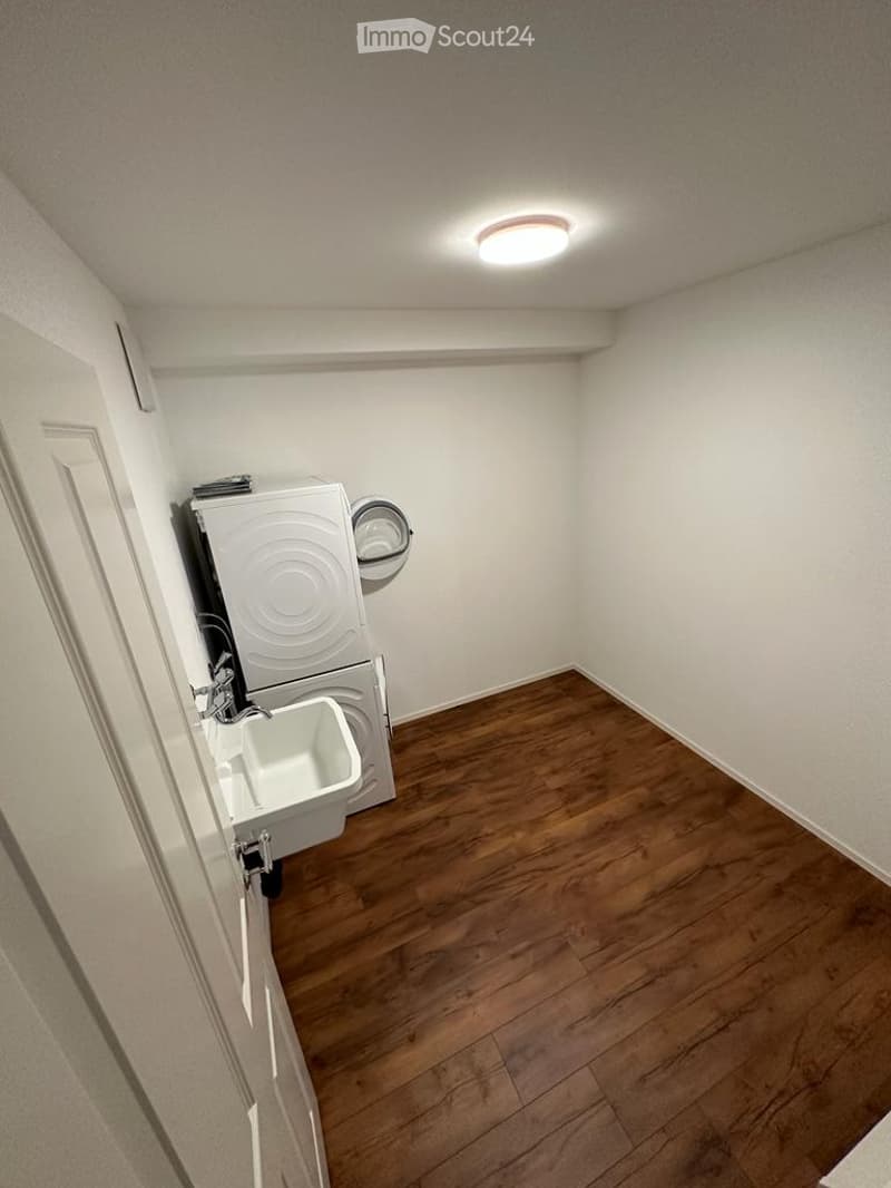 Small, unfurnished room with wooden flooring, white walls, and a private washing machine in the corner.
