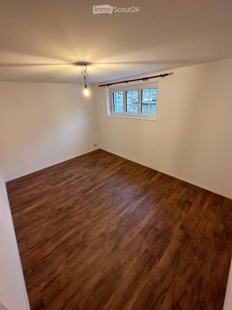 Completely empty room with wooden flooring, white walls, and a single window with white frame and black curtain rod.