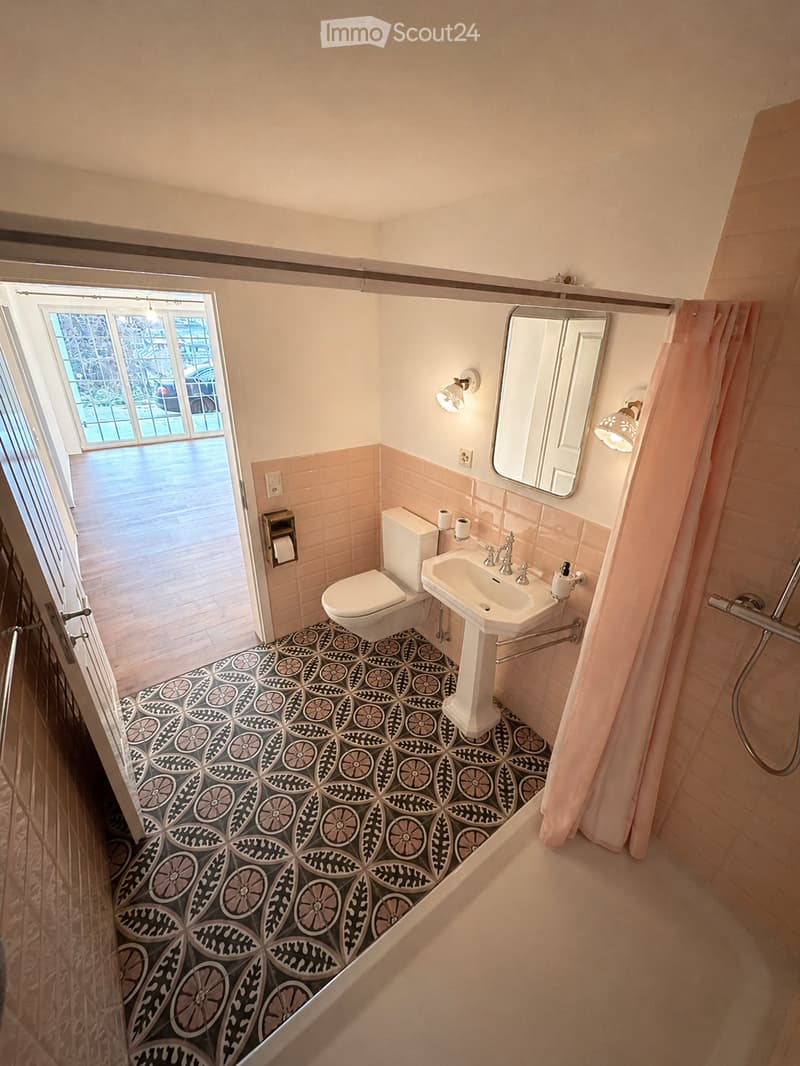 Bathroom with pink tiles, black and white patterned floor, toilet, sink, mirror, and shower area with a pink curtain.