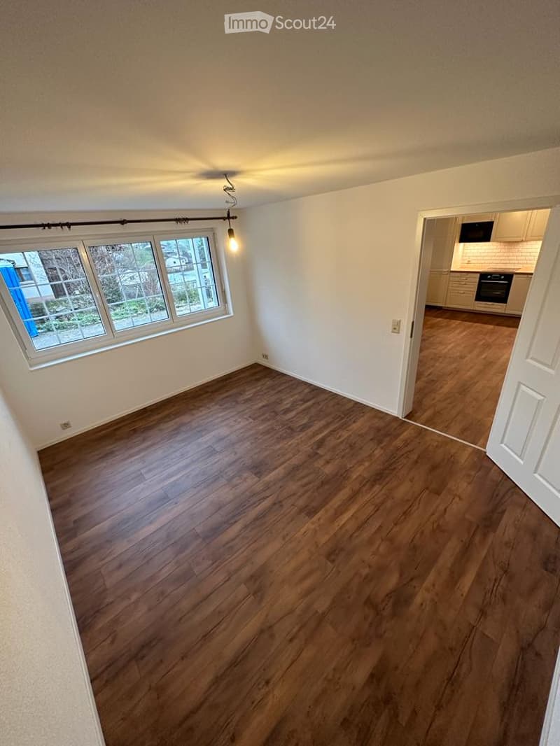 Empty room with wooden flooring, white walls, and a large window with blinds. Adjacent kitchen area visible through an open doorway.
