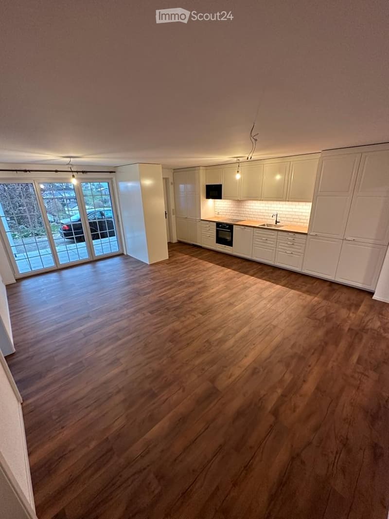 Open-plan space with wooden flooring, white kitchen cabinets, and a large glass sliding door leading to an outdoor area.