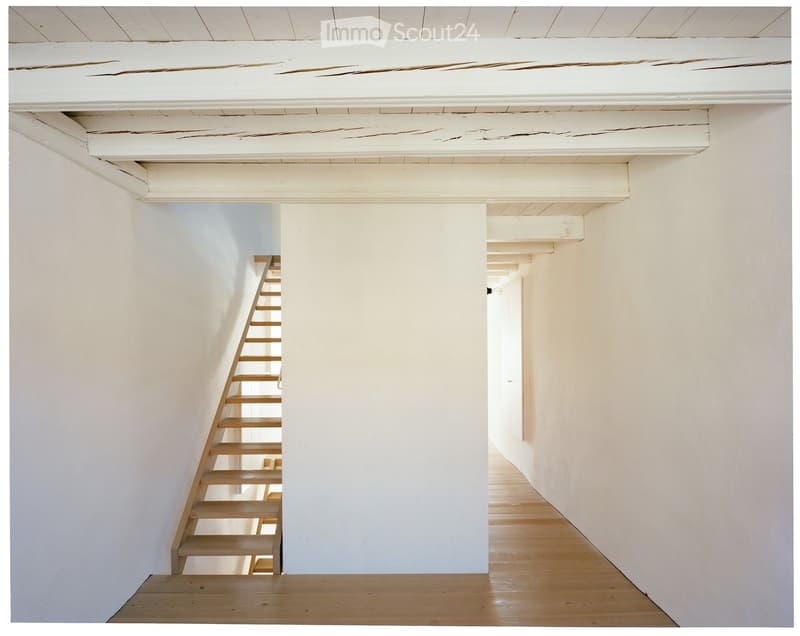 A narrow hallway with light wooden flooring and white walls, featuring a wooden staircase leading upwards and a white ceiling with visible beams and cracks.