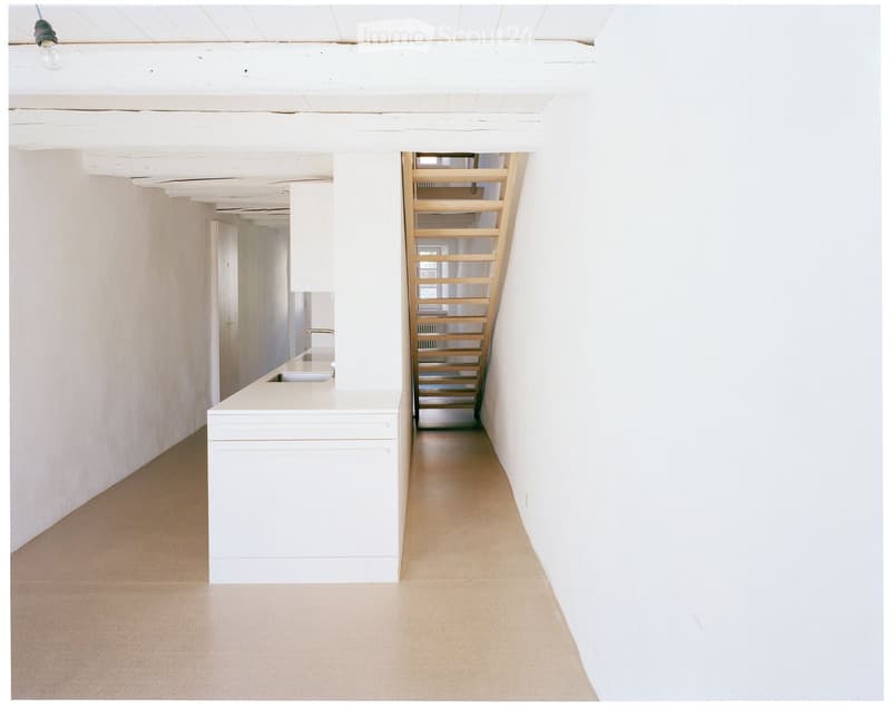 Minimalist hallway with white walls, light wooden staircase, and a small white counter with sink visible in the background.