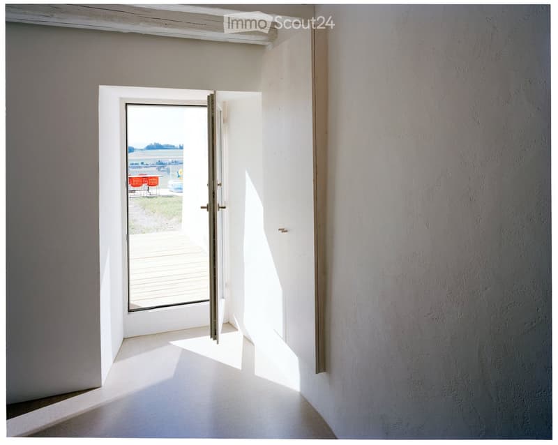 Bright, minimalist hallway with white walls and a large sliding glass door leading to an outdoor deck. Sunlight streams in, casting shadows on the floor.