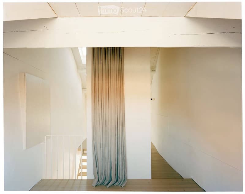 Bright, minimalist hallway with light-colored walls, wooden flooring, and a curtain partially covering a doorway. A white railing is visible on the left side, and a small staircase can be seen in the background.