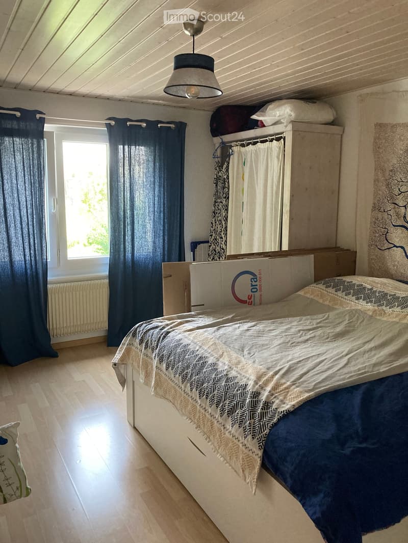 Bedroom with wooden flooring, a bed with patterned bedding, a wardrobe with hanging clothes, dark curtains, a radiator, and a mirror on the wall.