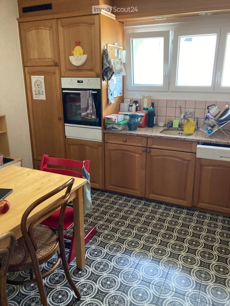 A cozy kitchen with wooden cabinets, a granite countertop, and a patterned tile floor. The space includes a wooden dining table with chairs, an oven, a sink, and various kitchen items on the counter.