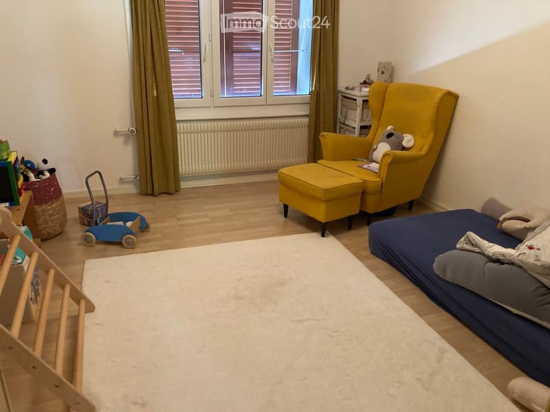 A cozy, well-lit room with a blue bed, yellow armchair, and a small wooden chair. The room features a white rug, a toy car, and a basket with toys.