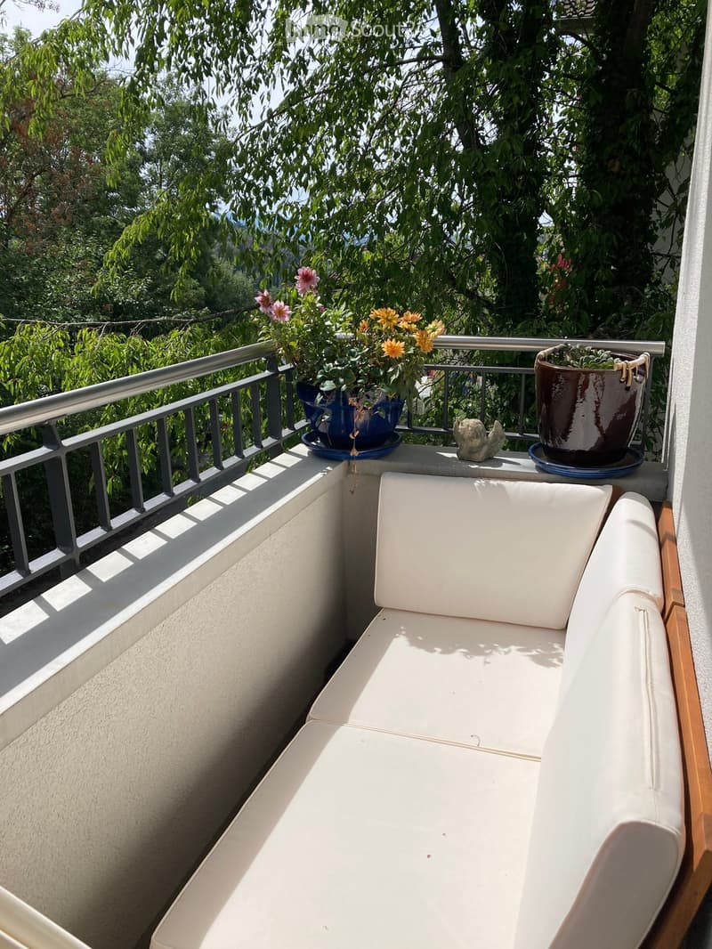 A balcony with a white leather sofa, flower pots, and a small statue, overlooking a green, leafy area.