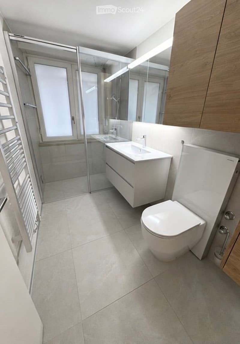 Modern bathroom with white tiles, a glass-enclosed shower, a floating sink, a toilet, and a large mirror with wooden cabinetry.