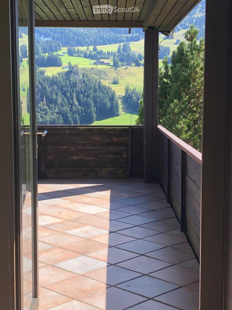 Balcony with tiled flooring and wooden railings, offering a scenic view of a green valley and forested hills.