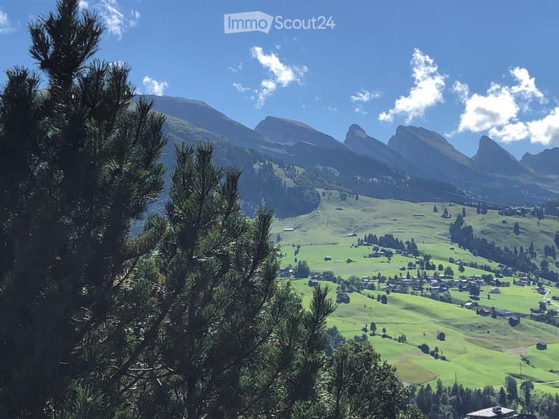 A panoramic view of a lush green valley with scattered houses, surrounded by towering mountains under a clear blue sky.