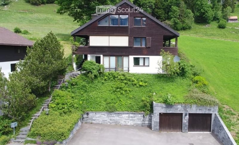 A two-story wooden house with a sloped roof, multiple windows, and a balcony on the upper level. The house is surrounded by greenery and has a stone-walled garage with two doors.