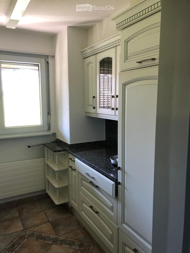 A small kitchen with white cabinets, a dark countertop, and a small bowl on the counter. There is a window with blinds and a small shelf unit next to the window.