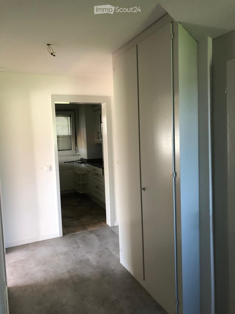 Empty hallway with beige walls, carpeted floor, and a partially open door leading to a kitchen area with cabinets and a countertop.