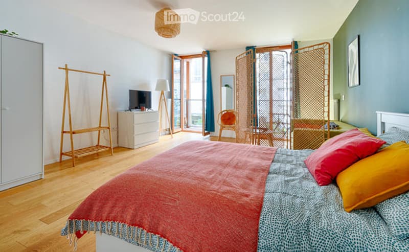 Bright bedroom with wooden floor, large windows, balcony access, bed with colorful blankets, dresser, chair, and screen.