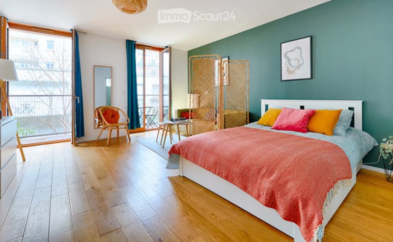 Bright bedroom with wooden flooring, large windows opening to a balcony, a neatly made bed with colorful pillows and a red blanket, a wooden chair, a small table, a mirror, and a decorative screen.