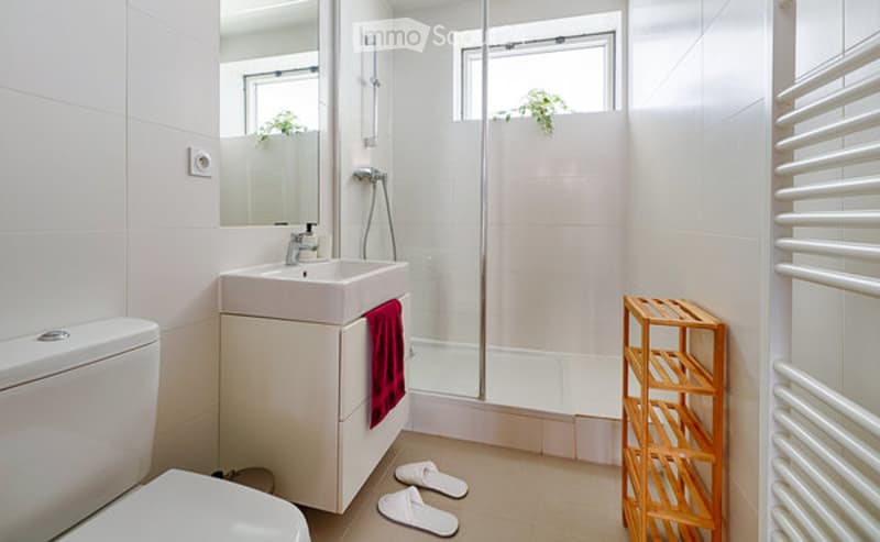 Modern bathroom with white tiles, a sink, mirror, toilet, glass-enclosed shower, a red towel, a wooden shelf, and two white bathroom slippers on the floor.
