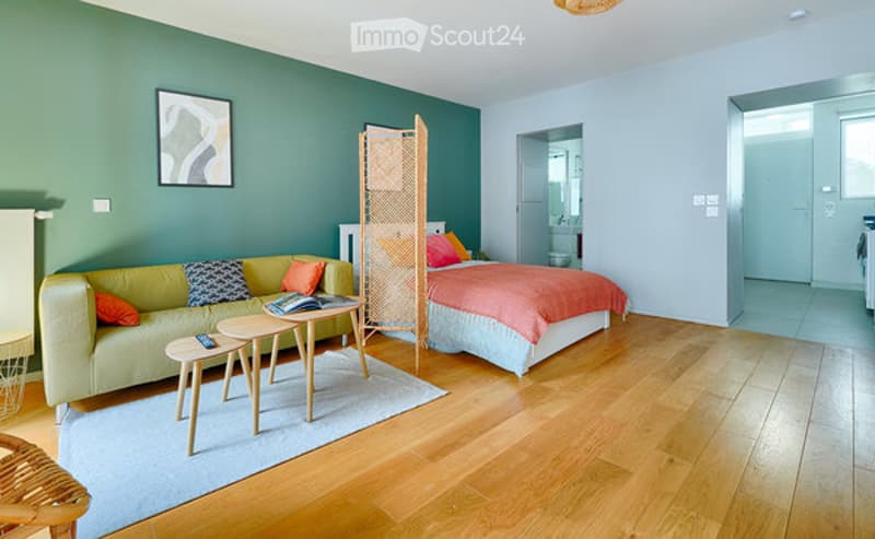 A modern, open-concept living and sleeping area with a green accent wall, a light green sofa with colorful pillows, a wooden bed with a pink and orange bedspread, a small wooden table, and a wicker chair. The room features hardwood flooring and a large wi