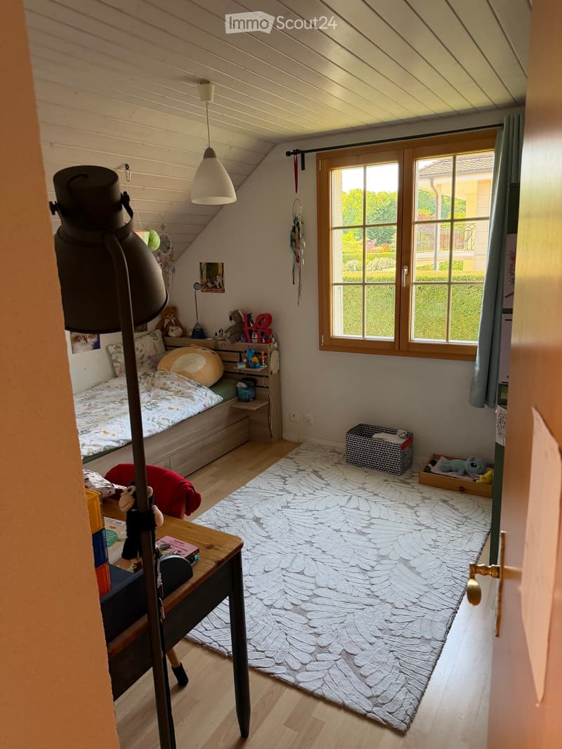 Cozy attic bedroom with slanted wooden ceiling, large window, bed with colorful bedding, small table, and various toys and decorations.