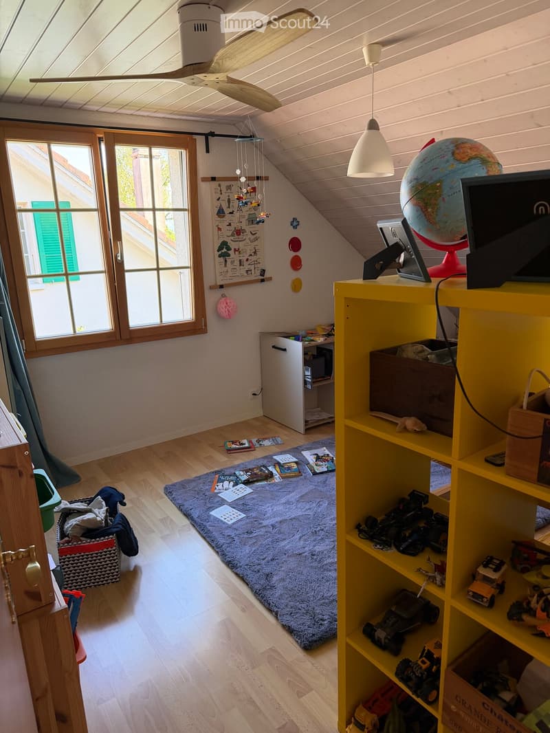 Brightly lit room with wooden floors, large windows, a yellow shelf filled with toys, a globe, a screen, and a small table with a chair. A blue rug and a hanging mobile add a playful touch.
